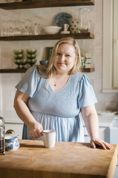 Woman in a light blue dress holding a coffee mug on the counter