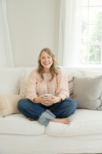 Woman in a pink blouse seated barefoot and criss-cross on a white couch holding a pink cell phone and looking up to smile