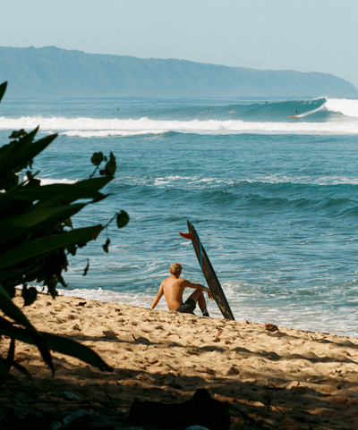 Young surfer on beach with board looking out at waves in Hawaii