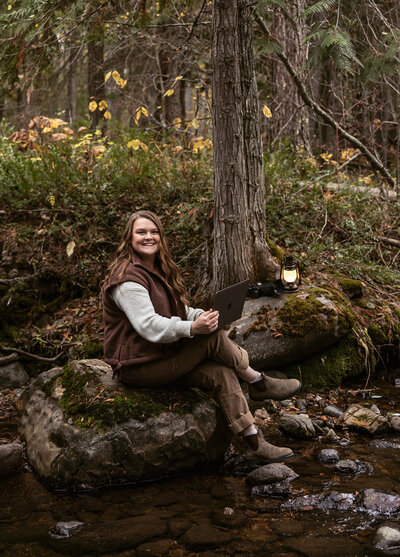 Sydney Breann sitting beside a creek in the forest, planning elopements on her laptop with a lantern and camera nearby, surrounded by autumn leaves.