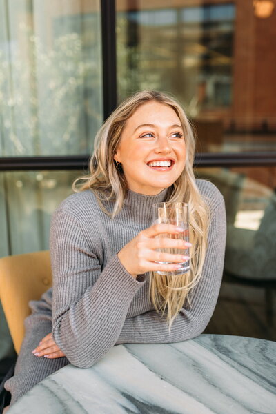 Iskra is sitting at a marble table holding a glass of water in one hand and smiling.