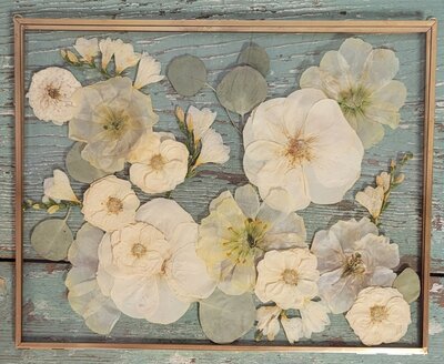Rustic pressed flower art featuring soft white and cream blooms with eucalyptus leaves, arranged in a gold frame over a distressed teal wooden backdrop.
