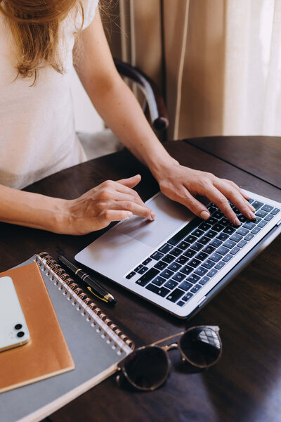 A female entrepreneur types on a laptop at a wooden table. Nearby are a smartphone, notebooks, a pen, and sunglasses, creating a focused and casual atmosphere.