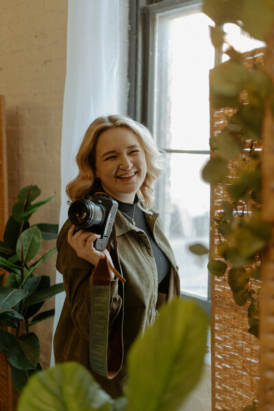 Woman smiling warmly, holding a camera by a window, surrounded by green plants. Sunlit room, cozy and inviting atmosphere.