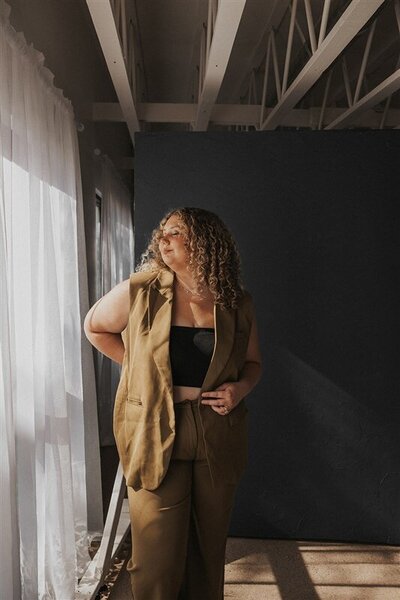 Andrea Cakmar, a wedding photographer, stands in a sunlit room, looking thoughtfully out the window. She is dressed in a tan pantsuit and black crop top, with soft natural light filtering through the sheer curtains.