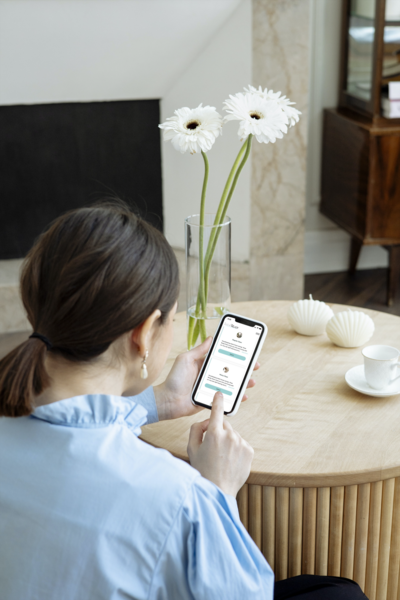 Woman in blue dress shirt scrolls her Iphone 11 cell phone. She sits in front of a circular wooden table with two white daisy flowers in a clear vase.