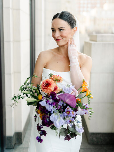 A white bride with dark hair poses with her bouquet, a splash of colorful purples and oranges.