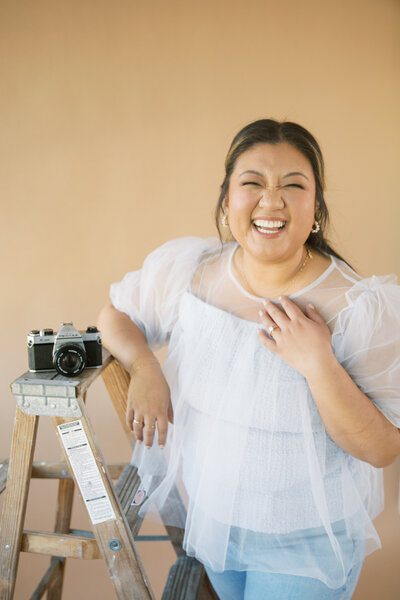 Monique Anne smiling next to a Pentax camera on a ladder