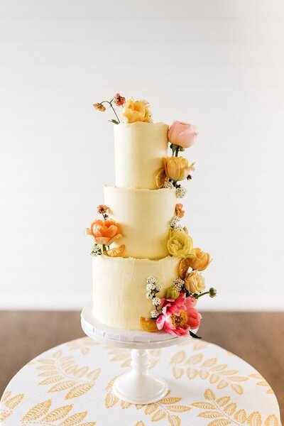 Photo shows a three tier wedding cake with pale yellow frosting, decorated down the side with a cascade of fresh flowers in pinks, yellows, and orange shades, with candied citrus.