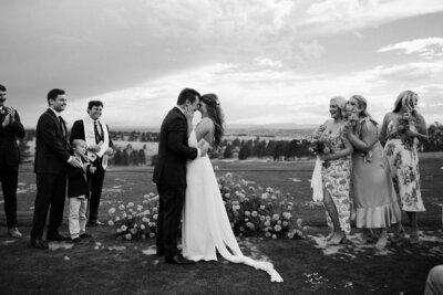 Newly weds stand together at the alter with their faces touching. They are surrounded by family with a flower bed and mountain scene in the background