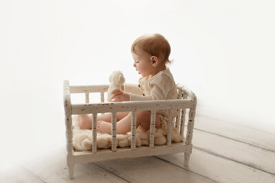 A young toddler in a white onesie sits in a wooden crib playing with some stuffed animals