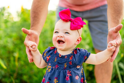 A baby wearing a bright pink bow and colorful dress giggles while her dad helps her walk.
