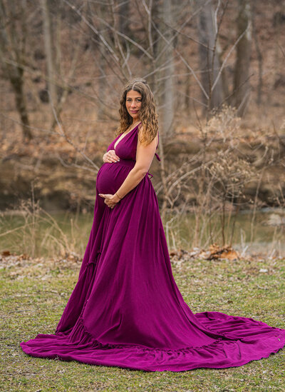 pregnant mom in purple dress standing outside holding belly and looking at camera