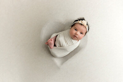 Baby girl with flower headband looks into camera while posing in a heart bowl.