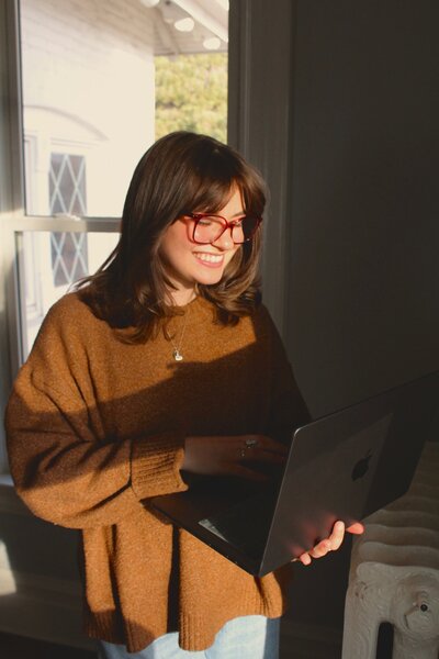 Woman with glasses smiling while typing on her laptop with one hand and holding the laptop with the other