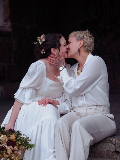 a newlywed eloping lesbian couple sit on stone steps and lean in to kiss, laughing and smiling. they both wear white, one in a dress holding a bouquet and one in a shirt and pants.