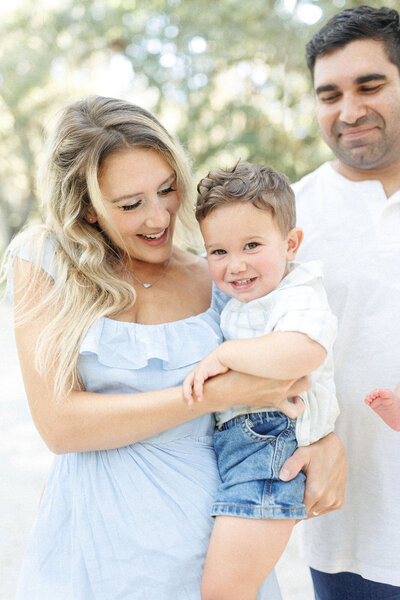 A mom in a blue dress holding her toddler son while dad watches smiling during their family photo session. 