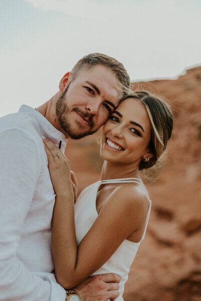 A close up shoot of a man and woman smiling at the camera. The woman has her hands on his chest with the wild landscape of Captiol Reef blurred in the background.