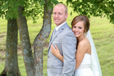 A white couple wearing wedding attire standing together. The bride is hugging the groom from behind