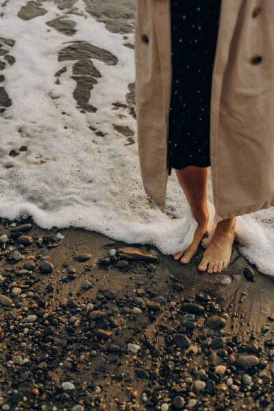 Frau, barfuß mit Kleid und Trenchcoat an der schaumigen Wasserkante stehend, vor ihr der kieselige Strand: Balance finden am Ufer des Lebens.