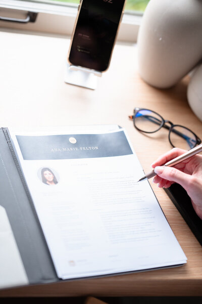A hand holding a pen reviews a printed document on a wooden desk. A pair of glasses, a smartphone on a stand, and ceramic vases are nearby.
