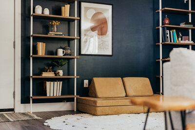 A lounge area featuring a tan seating cushions, wooden wall shelves decorated with books and ceramics, a framed abstract art piece, and a round white crocheted rug.
