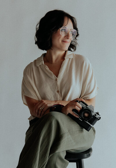 woman with glasses holding a camera, looking off to the side smiling.