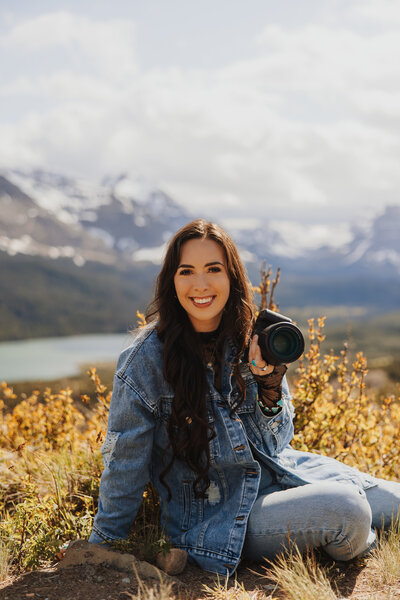 Aurelia, owner of Aurelia K Photography, smiling and holding her camera in front of a lake and mountain range in Glacier National Park.