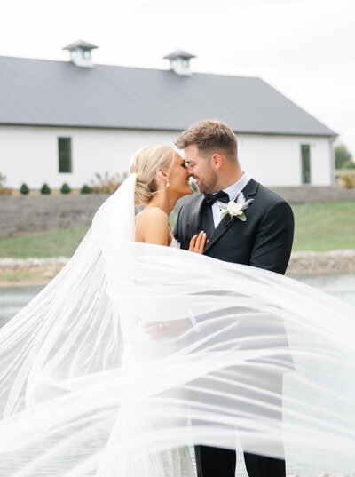 a bride with a long white veil and groom in black tuxedo kiss in front of a white barn