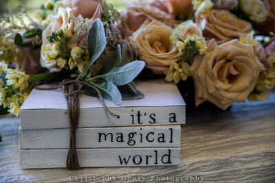 Wedding detail shot of stacked books with “It’s a magical world” text surrounded by peach roses