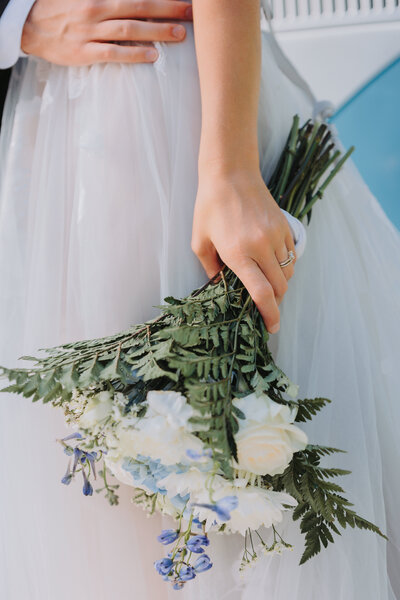 Bouquet of white and blue flowers being held in the left hand of a bride in a white dress with wedding ring highlighted