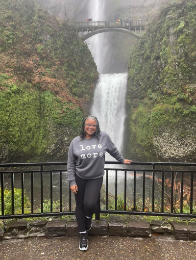 Letitia Hopkins standing confidently at an Oregon waterfall wearing a 'Love More' sweatshirt, embodying self-love, alignment, and Main Character Energy.