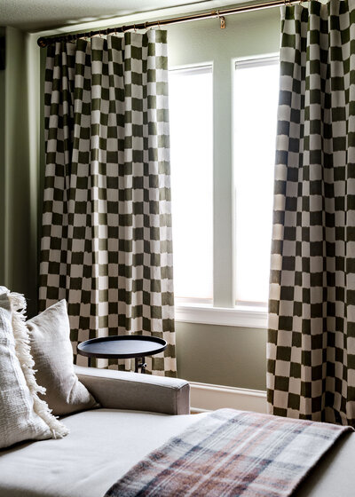 Corner of a room with a lounge chair  with a plaid blacker on it and checkered curtains over the window, representing a maximalist interior design.