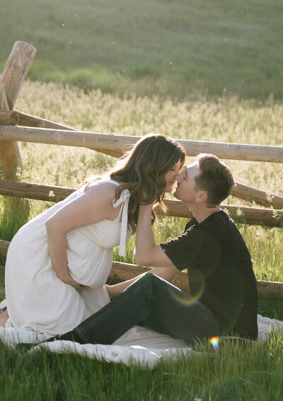 Expecting couple sharing a tender kiss in front of a rustic wooden fence during golden hour in Idaho