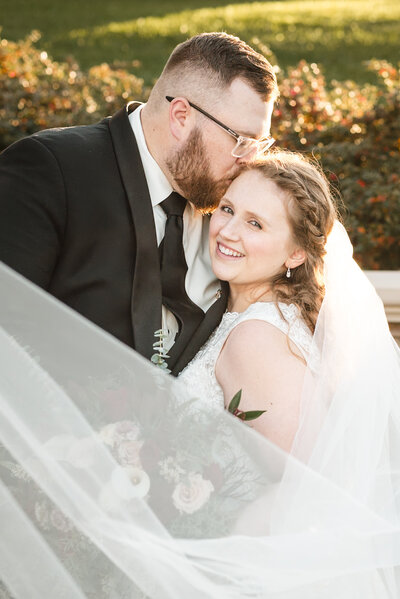 Groom kissing his bride's temple, as she smiles at the camera, holding a bouquet and her veil swooped around them to the foreground. The photograph was taken outside at Childress Vineyards in Lexington, NC.