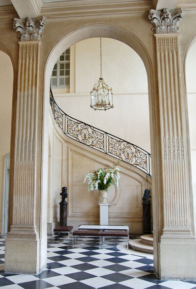 A grand staircase with ornate black railings curves upward in a classic foyer. Tall, fluted columns frame the scene, and a chandelier hangs above. A large floral arrangement sits atop a pedestal, and a checkered floor adds elegance.