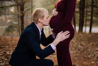 father in blue suit kisses the belly of his expectant wife who is wearing a burgandy fitted maternity gown in wooded location