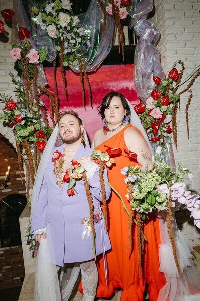 A queer couple stands in front of a colorful floral & fabric backdrop. One wears a lavender blazer dress and one wears a bright orange gown. Both wear veils and hold modern floral bouquets.