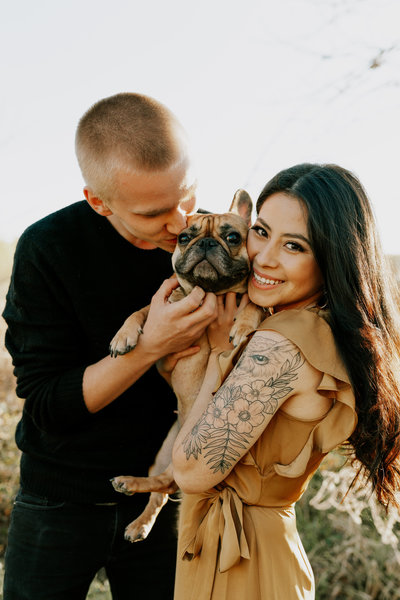 couple with tattoos holding and kissing their pug dog during summer engagement session in omaha nebraska, photo by photographer in omaha, Kaitlyn Neeley