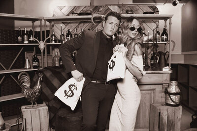 Couple posing in vintage photo booth with 1920s gangster props, dollar bags, lace umbrella, and retro bar backdrop by Philly Snap Booth