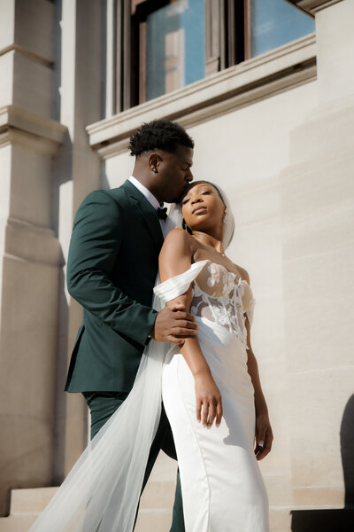 Couple embracing on courthouse steps, with the groom in a green suit and the bride in an off-the-shoulder lace gown with a flowing veil.