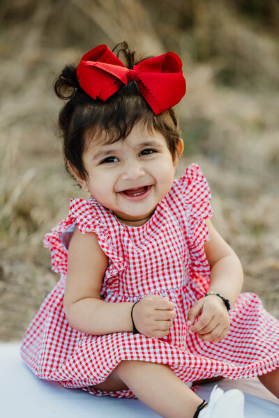 one year old baby portrait of little girl in red and white plaid dress and red bow sitting on white blanket in grass field