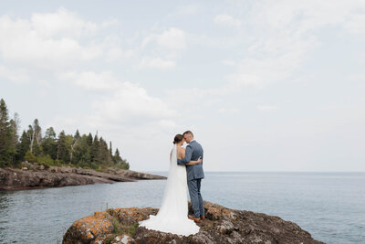 A newlywed couple standing head to head on a rock overlooking the water 