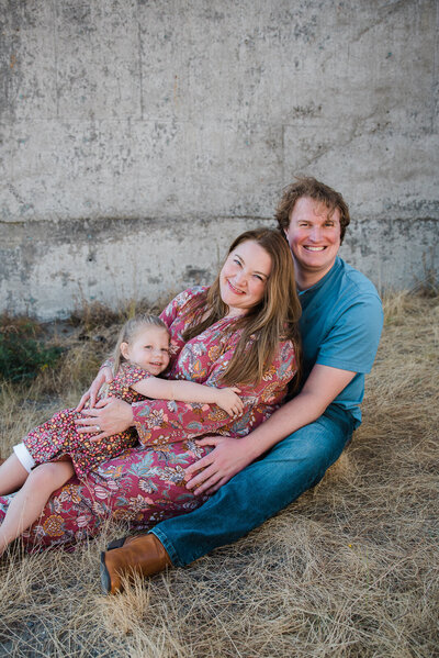 Family of 3 sitting on grass snuggled together mauve and blue outfit colors summer family photos