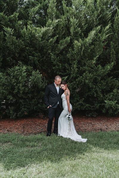 A newlywed couple shares a tender moment, embracing in front of a lush green backdrop of trees at The Gentry Farm in Danville, Virginia, captured beautifully by Quinn Photography LLC.
