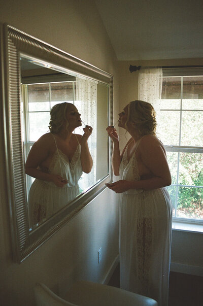 A bride puts lip gloss on while looking in a mirror, representing the documentary style of Oregon wedding photographer Among the Oaks. 