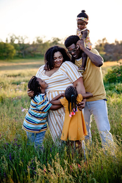 A family stands in a field, everyone showing affection towards someone else, and  Mom is in the middle of all of the hugs while Dad stands behind her with a child on his shoulders.