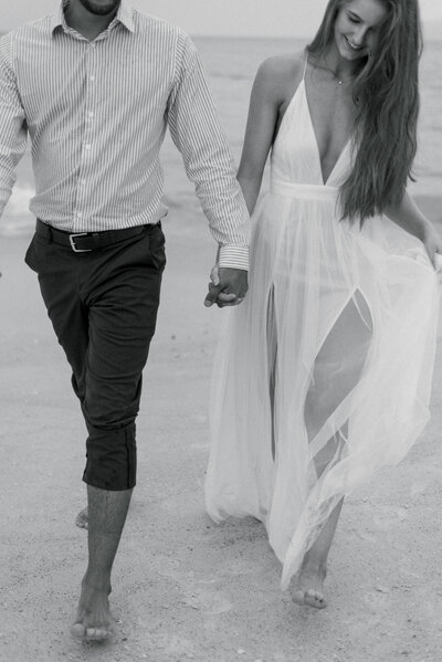 Black and white photo of a bride and groom walking hand in hand out of the water on St. Pete Beach in Florida bride wearing a long white halter dress with a thigh high slit