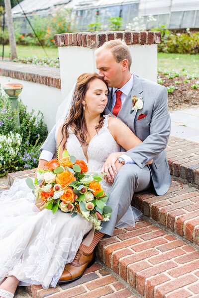 groom kissing brides head sitting down on steps at the Barn at Reynolda Village