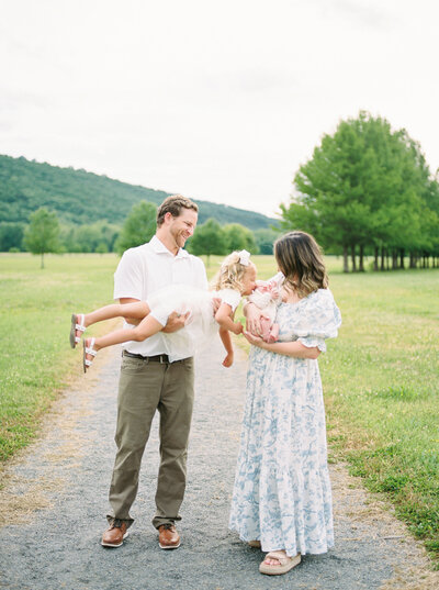Dad airplanes toddler daughter to her newborn baby sister being held by mother in a blue floral dress